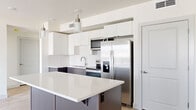 This well-appointed 2-bedroom, 2-bathroom residence offers a thoughtfully designed single-floor layout with a clean, contemporary aesthetic. The open-concept kitchen and living area serves as the heart of the home, featuring a large central island with a white quartz countertop, two-tone cabinetry in white and gray, stainless steel appliances including a refrigerator with ice dispenser, range, and microwave, all complemented by stylish pendant lighting. Light wood-look flooring runs throughout the main living areas, creating a bright, cohesive feel. The spacious living room is bathed in natural light from multiple windows fitted with blinds. The primary bedroom is generously sized, featuring soft carpet flooring, a large window with exterior views, and an en-suite bathroom that connects to a dedicated closet with built-in hanging rods. The primary bath includes a vanity with dark cabinetry and white countertops. The secondary bedroom also features carpet flooring, a double-door closet, and a bright window. The second bathroom offers a full tub-and-shower combination with a curved rod and matching dark vanity. The overall ambiance is bright, modern, and move-in ready, with a neutral palette that offers versatile styling potential.
