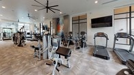 Located at 101 E Abram St in Arlington, Texas, this single-floor fitness center offers a comprehensive workout facility designed with both functionality and aesthetics in mind. The space is organized into distinct workout zones, each thoughtfully arranged to accommodate a wide range of exercise needs.

Upon entering, a clean, light-tiled entryway with a branded welcome mat leads into the main fitness area. The interior is finished with neutral-toned chevron-patterned carpeting throughout, contributing to a modern and cohesive visual identity. White walls and recessed lighting keep the space bright and open, while large black-framed windows along the perimeter allow ample natural light and outdoor views.

The cardio section features treadmills, ellipticals, stationary bikes, and other aerobic machines, complemented by wall-mounted flat-screen televisions for entertainment during workouts. Oversized ceiling fans ensure effective air circulation throughout. A dedicated strength training zone includes Hoist-brand resistance machines, a squat rack, free weights, and stability balls neatly displayed on a stand.

Mirrored walls in select areas visually expand the space and support form-checking during exercises. A built-in counter with cabinet storage provides a functional service station within the facility. The overall ambiance is clean, modern, and energizing, making it a well-rounded fitness destination.