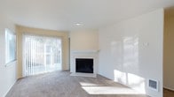 This single-floor space offers a clean, well-maintained interior designed for comfortable living. The main living area is the focal point of the space, centered around a classic white-mantled fireplace with a sleek black firebox surround — a standout architectural detail that adds warmth and character to the room. Soft, neutral-toned carpeting extends throughout the living area, providing a comfortable underfoot feel and complementing the light, airy color palette of warm white and cream-toned walls. Natural light plays a significant role in the ambiance of this space. A large sliding glass door fitted with vertical blinds allows generous amounts of daylight to flood the interior, creating a bright and welcoming atmosphere. The door also provides a view of the outdoor greenery beyond, adding a sense of connection to the outside. Recessed ceiling lighting supplements the natural light, ensuring the space remains well-lit throughout the day. The open layout of the living area offers flexibility for a variety of furniture arrangements, making it easy to personalize. The overall design is neutral and move-in ready, providing a versatile canvas for a range of interior styles.
