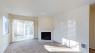 This single-floor space offers a clean, well-maintained interior designed for comfortable living. The main living area is the focal point of the space, centered around a classic white-mantled fireplace with a sleek black firebox surround — a standout architectural detail that adds warmth and character to the room. Soft, neutral-toned carpeting extends throughout the living area, providing a comfortable underfoot feel and complementing the light, airy color palette of warm white and cream-toned walls. Natural light plays a significant role in the ambiance of this space. A large sliding glass door fitted with vertical blinds allows generous amounts of daylight to flood the interior, creating a bright and welcoming atmosphere. The door also provides a view of the outdoor greenery beyond, adding a sense of connection to the outside. Recessed ceiling lighting supplements the natural light, ensuring the space remains well-lit throughout the day. The open layout of the living area offers flexibility for a variety of furniture arrangements, making it easy to personalize. The overall design is neutral and move-in ready, providing a versatile canvas for a range of interior styles.