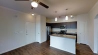 This single-floor residence features an open-concept layout that seamlessly connects the entryway, kitchen, and living area, creating a spacious and functional flow. The entryway is framed by crisp white doors and neutral-toned walls, establishing a clean, welcoming aesthetic upon arrival.

The kitchen is a standout feature, equipped with dark espresso cabinetry that extends to the upper walls, offering generous storage. A granite-topped peninsula serves as both a workspace and casual dining area, anchoring the kitchen within the open floor plan. Stainless steel appliances, including a refrigerator, electric range, and built-in microwave, provide a polished, modern look. A tile backsplash adds a subtle decorative element behind the range.

Overhead, a combination of pendant lights hanging above the peninsula and a track lighting system ensures ample task and ambient illumination throughout the kitchen. A ceiling fan with a light fixture serves the adjoining living area, enhancing comfort and airflow. Rich, dark hardwood-style flooring runs consistently throughout the space, tying the rooms together and adding warmth to the overall interior design.