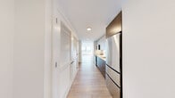 This well-appointed one-bedroom, one-bathroom residence offers a thoughtfully designed layout that maximizes every square foot of its compact footprint. Light hardwood flooring runs throughout the main living areas, complementing the clean, white walls and modern aesthetic that define the space.

The galley-style kitchen is sleek and functional, featuring dark lower cabinetry paired with white upper cabinets, stainless steel appliances including a full-sized refrigerator and oven, and a light countertop that runs the full length of the workspace. Louvered closet doors along the hallway provide convenient storage without interrupting the streamlined design.

The living area benefits from abundant natural light pouring in through a large picture window that frames expansive city views, creating a bright and airy atmosphere. The bedroom continues this connection to the outdoors with its own generously sized window offering similar scenic vistas. Carpeting in the bedroom adds warmth and comfort, and a set of double shaker-style closet doors provides ample storage.

The bathroom is finished to a high standard, featuring a wood-toned vanity with a white countertop, a large framed mirror with integrated medicine cabinet, tile surround, and a combined tub and shower. The overall design of the space is clean, contemporary, and move-in ready.