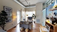 This single-floor urban loft blends industrial character with contemporary design, creating a stylish and functional living space. The open-concept living area immediately impresses with soaring ceilings that showcase exposed ductwork and concrete details, lending the space an authentic loft aesthetic. Warm hardwood flooring runs throughout, adding a natural contrast to the industrial overhead elements. Oversized windows flood the living area with abundant natural light, while sheer curtains provide a soft, layered look. A glass-panel door opens directly to a private balcony, extending the living space outdoors. The kitchen is thoughtfully outfitted with dark wood cabinetry, granite countertops, a mosaic tile backsplash, and black appliances including a full-size refrigerator and dishwasher, complemented by a built-in wine rack for added character. A striking full-wall cityscape mural anchors the transition between the kitchen and living zones, serving as a bold artistic focal point. The bathroom features bold teal and white horizontal striping, a combined tub and shower, a wood-toned vanity, and the convenience of a stacked in-unit washer and dryer. A lofted sleeping area accessible from within adds a creative vertical dimension to the layout.