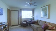 This single-floor space centers around a well-appointed living room that blends comfort and functionality. The room features carpeted flooring and neutral-toned walls that create a calm, welcoming atmosphere. A ceiling fan with a light fixture is installed overhead, providing both air circulation and ambient lighting. A wide, multi-pane window dressed with patterned curtains allows natural light to filter into the room, offering a view of the exterior surroundings. The living area is furnished with a upholstered sofa accented with decorative pillows and a matching armchair, arranged around a dark wood side table that holds a classic table lamp and a small potted plant. A framed mirror on the wall adds depth to the space. The front entry door, trimmed in warm wood tones, opens directly into the living area and is complemented by a welcome mat. The overall ambiance is relaxed and homey, with traditional furnishings and soft, muted tones contributing to a lived-in, comfortable feel throughout the space.