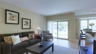 This single-floor residence features a comfortable and well-appointed living room as its central gathering space. The room is finished with light hardwood-style flooring and neutral beige walls, creating a clean, understated aesthetic throughout. A large sliding glass door dominates one wall, allowing abundant natural light to fill the space while providing direct views of lush greenery outside, enhancing the sense of openness and connection to the outdoors. Vertical blinds accompany the sliding door, offering flexible light control. The living area comfortably accommodates a dark upholstered sofa accented with decorative throw pillows, paired with a patterned armchair and a sleek black coffee table centered on a low-profile area rug. A framed piece of wall art and a classic table lamp contribute to the room's warm, inviting character. The layout is straightforward and functional, with clean sightlines and a smooth flow between the seating area and the access point to the outdoors. Overall, the space offers a calm, neutral environment well-suited for everyday living.