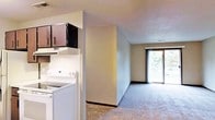 This single-floor residence offers a practical and comfortable layout designed for everyday living. The kitchen is equipped with a white electric range and features classic white tile backsplash, dark wood cabinetry overhead, and an integrated range hood, providing ample storage and a clean, functional workspace. The kitchen flows openly into the main living area, which is finished with wall-to-wall carpeting that adds warmth and softness underfoot. Neutral-toned walls throughout the space create a calm, understated atmosphere. A standout feature of the living area is the large sliding glass door at the far end, which floods the room with natural light and opens to a private balcony overlooking lush, mature trees. The balcony extends the living space outdoors, offering a pleasant retreat with greenery views. The overall design is straightforward and efficient, with a layout that maximizes usable space while maintaining an open, airy feel. The combination of natural light from the sliding glass door and the soft carpeted interiors gives the space a welcoming and livable ambiance suited to a variety of lifestyles.