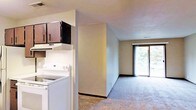 This single-floor residence offers a practical and comfortable layout designed for everyday living. The kitchen is equipped with a white electric range and features classic white tile backsplash, dark wood cabinetry overhead, and an integrated range hood, providing ample storage and a clean, functional workspace. The kitchen flows openly into the main living area, which is finished with wall-to-wall carpeting that adds warmth and softness underfoot. Neutral-toned walls throughout the space create a calm, understated atmosphere. A standout feature of the living area is the large sliding glass door at the far end, which floods the room with natural light and opens to a private balcony overlooking lush, mature trees. The balcony extends the living space outdoors, offering a pleasant retreat with greenery views. The overall design is straightforward and efficient, with a layout that maximizes usable space while maintaining an open, airy feel. The combination of natural light from the sliding glass door and the soft carpeted interiors gives the space a welcoming and livable ambiance suited to a variety of lifestyles.