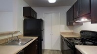 This single-floor residence offers a practical and well-organized layout designed for comfortable everyday living. The galley-style kitchen is efficiently arranged with dark espresso cabinetry lining both sides, providing ample upper and lower storage. Black appliances, including a refrigerator and a coil-top electric range, complement the dark cabinetry, while light-toned countertops offer a subtle contrast. A stainless steel sink is conveniently positioned along one wall, and a set of bi-fold doors leads to additional pantry or utility storage at the end of the kitchen corridor. Wood-look flooring grounds the kitchen space with a warm, durable finish. The living area transitions to plush carpeting, creating a cozy and inviting atmosphere. A ceiling fan with an integrated light fixture serves as the central feature of the room, providing both air circulation and ambient lighting. A peninsula counter connects the kitchen to the living space, offering a natural division between the two areas. Track lighting adds supplemental illumination along one wall, and a doorway leads directly into the bedroom beyond. The overall layout is straightforward and functional, making efficient use of the available space throughout.