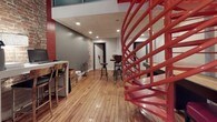 This single-floor commercial space masterfully combines industrial character with modern functionality. The exposed brick walls serve as a defining architectural feature, lending warmth and texture throughout the space. Warm-toned hardwood floors run continuously underfoot, complementing the raw aesthetic and tying the various areas together cohesively.

A striking red spiral staircase commands attention as a central design element, acting as both a sculptural focal point and a functional architectural feature. The open layout allows for multiple distinct work and activity zones. A white counter-height workstation is equipped with bar-style seating and features built-in workspace for computers and office equipment, making it ideal for collaborative tasks or administrative work.

A recreational area includes a shuffleboard table accompanied by casual seating, offering a social or breakroom-style environment. Industrial-style adjustable bar stools are positioned throughout, reinforcing the loft-like character of the space. Recessed lighting in a drop-tile ceiling section provides even illumination alongside arced floor lamps that add a softer ambiance near the workstations.

Storage shelving units provide practical organization near the entry area, and a round wall clock adds a utilitarian touch. The overall atmosphere is creative, energetic, and purpose-built for a dynamic professional or co-working environment.