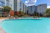 Located at 800 W Marietta St NW in Atlanta's vibrant Westside neighborhood, this outdoor amenity space offers a well-appointed, resort-inspired environment designed for relaxation and social gatherings. At the heart of the space is a generously sized swimming pool, surrounded by rows of sleek lounge chairs that invite residents to unwind under the open sky. The pool deck is finished in a textured aggregate concrete surface, providing both durability and visual appeal.

A standout architectural feature is the modern steel-and-wood pergola, which anchors a dedicated lounge and bar-height seating area finished with a bold green rubberized surface. Vibrant yellow bar chairs and dark high-top tables create a lively, social atmosphere beneath the open-slatted wood canopy. Additional high-top seating is available on the main deck, offering flexible gathering options.

The space is framed by mature trees, cypress columns, ornamental shrubs, and curated landscaping beds that provide natural privacy and a lush, green aesthetic. A built-in outdoor grilling station, featuring stainless steel grills set into a brick-clad wall, adds a functional and inviting element for outdoor cooking. The surrounding brick and siding building architecture adds an urban, contemporary character to the overall environment.