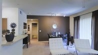 This well-appointed single-floor space showcases a thoughtful blend of modern design and functional living. The open-plan layout seamlessly connects the living and dining areas, creating a cohesive and inviting atmosphere. A striking dark-painted accent wall serves as the focal point of the room, adorned with a decorative sunburst mirror that adds a glamorous touch. Track lighting runs along the ceiling, providing targeted illumination that highlights the space's key features.

The living area is furnished with sleek, contemporary seating, including a white tufted chair with chrome legs that adds a sophisticated contrast to the darker tones of the room. The dining area accommodates a dark table with matching chairs, complemented by elegant décor pieces including tall vases and a side table. Soft, neutral-toned carpeting flows throughout, adding warmth underfoot.

A walk-in closet is conveniently accessible from the main living space, offering generous storage with visible hanging rails. The overall ambiance is polished and modern, with a carefully curated color palette of deep charcoal, crisp white, and warm neutrals that creates a balanced and stylish interior environment.