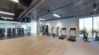 This well-appointed fitness facility combines industrial aesthetics with functional design to create an inviting and practical workout environment. The space is defined by its striking exposed concrete ceiling, which features visible ductwork and structural elements, lending the room an urban, loft-like character. Suspended linear fluorescent lighting and track lighting provide bright, even illumination throughout the space.

The flooring transitions between warm hardwood wood panels in the open exercise area and rubberized matting near the equipment zones, offering both visual appeal and practical support underfoot. Along one wall, a lineup of modern cardio machines — including treadmills and elliptical trainers — are equipped with individual digital screens, catering to connected, technology-driven workouts.

A multi-functional cable and weight machine anchors the strength training section, while a dedicated free weights area with a dumbbell rack and additional equipment is visible toward the rear. Large mirrors along the far wall enhance the sense of depth and allow users to monitor their form. The open floor plan provides generous space for stretching or floor-based exercises, making this facility versatile and well-suited for a wide range of fitness routines.