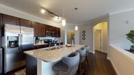 This single-story residence at 9815 Seasons West Dr. in Indianapolis offers a thoughtfully designed layout with refined finishes throughout. The kitchen is a standout feature, boasting rich dark cabinetry, granite countertops, a subway tile backsplash, stainless steel appliances, and a large center island with an undermount sink and seating for multiple barstools — ideal for casual dining and entertaining. Crown molding and dark hardwood-style flooring flow throughout the main living areas, adding a polished, cohesive aesthetic. The dedicated dining area accommodates a round table and features a classic pendant light fixture, creating an intimate setting. The living room is bright and inviting, with neutral walls and crown molding that lend an airy, comfortable atmosphere. Two well-sized bedrooms offer carpeted floors and ample natural light from large windows; one features a ceiling fan for added comfort. The bathroom includes a tub-shower combination with classic white subway tile, a dark wood vanity with granite countertops, and hardwood-style flooring. A dedicated in-unit laundry room with a full-size washer and dryer adds everyday convenience. A walk-in closet with built-in shelving provides generous storage. The overall ambiance is clean, modern, and functional.