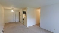 This single-floor space features an open-concept layout that connects the living area to the kitchen through a thoughtful partial wall divider, creating a sense of separation while maintaining an open feel. The living area is finished with neutral-toned wall-to-wall carpeting, contributing to a warm and understated ambiance throughout the space.

The kitchen is visible from the main living area and is equipped with a white electric range and overhead range hood, complemented by dark-toned upper cabinetry. The under-cabinet lighting provides functional illumination for the cooking area. A textured ceiling runs throughout the unit, adding subtle visual interest to the otherwise clean and straightforward design.

Near the entryway, a pair of closet doors offers practical storage solutions, while the entry door itself features a standard residential design. The neutral wall tones and light carpeting create a cohesive and functional interior that is easy to personalize. Overall, the space presents a straightforward, practical layout well-suited for everyday living, with distinct zones for the living and kitchen areas defined by the central dividing wall.