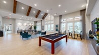 Located at 19025 Stuebner Airline Road in Spring, TX, this single-floor community gathering space offers a thoughtfully designed open-plan layout combining a living room and dining area in one cohesive environment. The space immediately impresses with its dramatic vaulted ceilings adorned with rich, dark wood exposed beams, complemented by recessed lighting and large pendant fixtures that create a warm, welcoming ambiance.

Light hardwood flooring flows throughout, tying the spaces together seamlessly. Floor-to-ceiling windows and a grand window wall flood the interior with abundant natural light while offering views of the surrounding community. The living area features stylish teal accent chairs arranged around a central coffee table, anchored by a patterned area rug, with a large wall-mounted television set against a shiplap accent wall.

A full-size pool table serves as a standout recreational feature, adding a social, leisure-oriented character to the space. The dining area accommodates a large communal table with woven rattan chairs, illuminated by a cluster of industrial-style pendant lights. Adjacent to the dining area is a kitchenette with navy cabinetry, quartz countertops, a sink, and built-in appliances, making the space well-equipped for gatherings and events. Overall, the space balances modern design with casual comfort.
