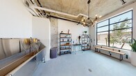 Located at 3725 N 27th St in Tacoma, WA, this single-floor pet amenity space blends functional design with modern industrial aesthetics. The open layout features exposed concrete columns and ceiling with visible ductwork and piping, lending an urban loft character to the room. A striking multi-bulb chandelier with an industrial pipe frame serves as a focal point, casting warm light throughout the space.

The walls are finished in white with a distinctive decorative tile wainscoting accented by colorful rectangular tiles in navy, teal, and gray, adding a playful yet polished look. Large, black-framed windows flood the room with natural light and offer views of the surrounding greenery outside.

A professional-grade stainless steel pet washing station occupies one corner, complete with a handheld sprayer and non-slip step stool for easy access. Adjacent to it is a grooming table equipped with a mounted dryer arm, ideal for post-wash care. Open metal and wood shelving units display grooming supplies and accessories in an organized, boutique-style arrangement.

An indoor pet relief area with an artificial turf insert and a decorative fire hydrant provides a convenient solution for pets. A live-edge wood bench offers seating for owners. A bold black-and-white photographic artwork of two bulldogs adds a charming finishing touch to this well-appointed space.