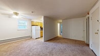 This single-floor residence offers a straightforward, functional layout with distinct living, sleeping, kitchen, and bathroom areas. The spacious living area is finished with neutral-toned wall-to-wall carpeting and features a ceiling fan, a baseboard heating system, and a window unit for cooling. The open sightline to the kitchen creates a connected feel between the two spaces.

The kitchen is equipped with warm honey-toned wood cabinetry, a textured tile backsplash, gray countertops, and white appliances including a gas range, refrigerator, and an overhead ventilation hood. Light tile flooring completes the kitchen's clean, practical aesthetic.

The bedroom is a generously sized room featuring two windows that bring in natural light and views of outdoor greenery, along with baseboard heating and carpeted flooring. An additional room adjacent to the living area, also carpeted and windowed, provides further flexible living space.

The bathroom is finished with a white tub-shower combination, a toilet, and a distinctive mosaic tile floor featuring a teal grid pattern, lending a classic character to the space. Throughout the unit, neutral white walls and consistent carpeting create a cohesive, understated atmosphere.