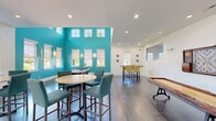 Located at Walkertown Landing in Walkertown, NC, this single-floor community game and recreation room offers a well-appointed, open-concept layout designed for social gatherings and leisure. The space is immediately distinguished by a striking accent wall finished in bold teal, which rises to double-height ceilings lined with clerestory windows that flood the interior with natural light. Polished concrete floors run throughout, lending an industrial-modern aesthetic that complements the clean white walls and recessed ceiling lighting. The game area is anchored by a full-size pool table and a classic shuffleboard table with vintage-style cast iron legs, providing ample entertainment options. A wall-mounted flat-screen television and a decorative board game display add additional focal points. Multiple high-top bistro tables paired with teal upholstered bar chairs are arranged throughout, offering casual seating for groups of various sizes. A separate dining-height table with yellow-green chairs provides an alternate gathering spot. Entry is through a set of modern glass and aluminum doors that connect to a polished lobby area, enhancing the sense of openness. Windows along multiple walls ensure natural light reaches every corner, creating a bright, welcoming atmosphere ideal for community interaction.