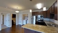 This single-floor residence offers a thoughtfully designed open-concept layout that seamlessly connects the kitchen and living areas. The kitchen is a standout feature, showcasing rich dark wood cabinetry that contrasts elegantly with light granite countertops. Stainless steel appliances — including a refrigerator, electric range, and built-in microwave — lend a modern, polished look. A kitchen island with a generous granite surface provides additional prep space and serves as a natural gathering point. Pendant lighting hangs above, adding warmth and character to the space. Durable hardwood flooring runs throughout, complementing the neutral wall tones and creating a cohesive, inviting atmosphere. A dedicated in-unit laundry area is conveniently accessible just off the main living space, housing a full-sized washer and dryer. A bathroom is also visible, fitted with standard fixtures. Vaulted ceilings enhance the sense of openness and airiness throughout. The overall design balances practicality with aesthetic appeal, making it a functional and comfortable living environment.