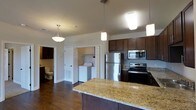 This single-floor residence offers a thoughtfully designed open-concept layout that seamlessly connects the kitchen and living areas. The kitchen is a standout feature, showcasing rich dark wood cabinetry that contrasts elegantly with light granite countertops. Stainless steel appliances — including a refrigerator, electric range, and built-in microwave — lend a modern, polished look. A kitchen island with a generous granite surface provides additional prep space and serves as a natural gathering point. Pendant lighting hangs above, adding warmth and character to the space. Durable hardwood flooring runs throughout, complementing the neutral wall tones and creating a cohesive, inviting atmosphere. A dedicated in-unit laundry area is conveniently accessible just off the main living space, housing a full-sized washer and dryer. A bathroom is also visible, fitted with standard fixtures. Vaulted ceilings enhance the sense of openness and airiness throughout. The overall design balances practicality with aesthetic appeal, making it a functional and comfortable living environment.