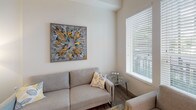 This single-floor unit showcases a clean, modern design with an open-concept layout that seamlessly connects the main living areas. The kitchen is a standout feature, outfitted with flat-front white cabinetry extending to the ceiling, a textured stone tile backsplash, stainless steel appliances including a range, built-in microwave, and dishwasher, and a white quartz peninsula with seating. Open shelving adds both function and visual interest to the cooking space, while a large window allows natural light to filter in.

The living area is bright and inviting, featuring wood-look flooring, neutral walls, and multiple windows dressed with white horizontal blinds that offer views of surrounding greenery. The space is well-proportioned, providing comfortable room for seating arrangements.

A dedicated in-unit laundry area with a stacked washer and dryer adds everyday convenience. The bathroom is finished with large-format gray tile flooring, a tub-shower combination with full tile surround, and a clean vanity setup, maintaining the unit's consistent modern aesthetic. Throughout the space, recessed lighting, high ceilings, and a cohesive neutral palette create a bright, contemporary atmosphere.