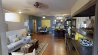 This single-floor residence showcases a thoughtfully designed open-concept layout that connects the living, dining, and kitchen areas into one cohesive and inviting space. Rich hardwood flooring flows throughout, anchoring the bold interior palette of deep charcoal-gray walls accented with vibrant yellow décor touches that create a modern and energetic ambiance.

The living area is furnished with a comfortable seating arrangement centered around a distinctive natural wood coffee table, complemented by a patterned area rug. An open shelving unit provides both display and storage, adding an industrial design element to the space. A ceiling fan overhead ensures comfort throughout.

The dining area features a round table with upholstered chairs positioned beneath a pendant light fixture, set against a striking yellow statement artwork that serves as a bold focal point on the gray accent wall.

The kitchen is well-appointed with a breakfast bar fitted with bar-height seating, stone countertops, warm wood cabinetry, and a track lighting system overhead. A wine rack and pendant lighting above the bar area add character and functionality. The open design of the kitchen allows it to interact naturally with the rest of the living space, making it ideal for entertaining and everyday living.