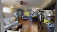 This single-floor residence showcases a thoughtfully designed open-concept layout that connects the living, dining, and kitchen areas into one cohesive and inviting space. Rich hardwood flooring flows throughout, anchoring the bold interior palette of deep charcoal-gray walls accented with vibrant yellow décor touches that create a modern and energetic ambiance.

The living area is furnished with a comfortable seating arrangement centered around a distinctive natural wood coffee table, complemented by a patterned area rug. An open shelving unit provides both display and storage, adding an industrial design element to the space. A ceiling fan overhead ensures comfort throughout.

The dining area features a round table with upholstered chairs positioned beneath a pendant light fixture, set against a striking yellow statement artwork that serves as a bold focal point on the gray accent wall.

The kitchen is well-appointed with a breakfast bar fitted with bar-height seating, stone countertops, warm wood cabinetry, and a track lighting system overhead. A wine rack and pendant lighting above the bar area add character and functionality. The open design of the kitchen allows it to interact naturally with the rest of the living space, making it ideal for entertaining and everyday living.
