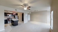 This single-floor residence offers an open-concept layout that seamlessly connects the living area and kitchen, creating a bright and functional living environment. The generously sized living area is finished with plush wall-to-wall carpeting, providing a comfortable and inviting atmosphere. A ceiling fan with a built-in light fixture is centrally positioned overhead, complemented by a track lighting system that adds both practicality and a modern touch to the space. The kitchen is well-appointed with dark espresso cabinetry, a contrasting light-toned countertop island that doubles as a breakfast bar, and stainless steel appliances including a refrigerator. Decorative pendant lights in deep red hang above the kitchen area, adding a warm accent and visual interest. A full-size black refrigerator is also visible, suggesting ample storage capacity. The transition between the kitchen and living space is defined by a partial half-wall, maintaining an open feel while providing subtle zoning. Neutral warm-beige walls throughout contribute to a cohesive and clean aesthetic. A white interior door leads to additional areas of the unit, rounding out this well-designed, move-in ready space.