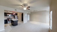 This single-floor residence offers an open-concept layout that seamlessly connects the living area and kitchen, creating a bright and functional living environment. The generously sized living area is finished with plush wall-to-wall carpeting, providing a comfortable and inviting atmosphere. A ceiling fan with a built-in light fixture is centrally positioned overhead, complemented by a track lighting system that adds both practicality and a modern touch to the space. The kitchen is well-appointed with dark espresso cabinetry, a contrasting light-toned countertop island that doubles as a breakfast bar, and stainless steel appliances including a refrigerator. Decorative pendant lights in deep red hang above the kitchen area, adding a warm accent and visual interest. A full-size black refrigerator is also visible, suggesting ample storage capacity. The transition between the kitchen and living space is defined by a partial half-wall, maintaining an open feel while providing subtle zoning. Neutral warm-beige walls throughout contribute to a cohesive and clean aesthetic. A white interior door leads to additional areas of the unit, rounding out this well-designed, move-in ready space.