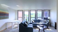 This single-floor space centers around a well-appointed living area designed for both comfort and functionality. The living room is furnished with a coordinated set of blue-grey sofas accented with patterned throw pillows, arranged around a sleek glass-top coffee table that adds a contemporary touch. A compact white desk and chair are positioned near the wall, offering a practical workspace within the living area.

A defining feature of this space is the row of large windows spanning the length of the room, fitted with dark-framed blinds that allow for light control. These windows flood the interior with generous natural light and provide elevated views of surrounding greenery and neighboring brick structures, creating a pleasant indoor-outdoor connection. The neutral-toned carpet and white walls contribute to a clean, airy atmosphere throughout.

The overall ambiance is bright, modern, and inviting, with a layout that balances open living space with thoughtful furnishing. The abundance of natural light and tree-lined views are standout qualities that enhance the livability and appeal of this space.