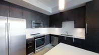 This single-level kitchen showcases a contemporary design defined by a bold contrast between dark espresso cabinetry and bright white countertops and backsplash. The layout follows an efficient L-shaped configuration, maximizing workspace and storage throughout. Floor-to-ceiling upper cabinets with sleek bar-style hardware provide generous storage capacity, while lower cabinets and drawers mirror the same refined aesthetic. Stainless steel appliances, including a slide-in electric range, built-in microwave, dishwasher, and a full-sized refrigerator, are seamlessly integrated into the cabinetry for a cohesive, streamlined look. A stainless steel undermount sink with a modern faucet is positioned along the perimeter counter, complementing the overall design palette. The white backsplash extends cleanly between upper and lower cabinets, enhancing the sense of brightness in the space. A recessed ceiling detail frames a cylindrical pendant light above, adding a focused architectural element overhead. Light-toned flooring grounds the space and provides a subtle contrast to the dark cabinetry. A white kitchen island or countertop is partially visible in the foreground, suggesting additional workspace and potential for casual dining or food preparation.