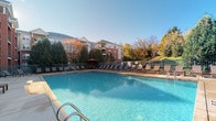 This amenity facility serves as a recreational hub for its surrounding residential community, offering a thoughtfully designed combination of indoor and outdoor spaces. The centerpiece is a generously sized outdoor swimming pool with crystal-clear blue water, surrounded by a wide pool deck furnished with rows of lounge chairs — ideal for relaxing under open skies. The pool area is enclosed by a green metal fence and is bordered by mature evergreen and deciduous trees, providing natural privacy and scenic views of the surrounding landscaped hillside. A multi-story brick residential building is visible beyond the tree line, reinforcing the community-oriented setting.

Inside, a dedicated pool house provides convenient access to the outdoor area through wood-framed glass doors that flood the corridor with natural light. The interior hallway features tile flooring and connects to key amenities, including a glassed-in hot tub room with mosaic tile finishes and posted safety guidelines. A row of lockers lines the hallway, offering secure storage for residents. A vending machine and decorative indoor plant near the entrance add a practical, welcoming touch to the facility. Large windows within the indoor space frame direct views out to the pool deck, maintaining a visual connection between the interior and outdoor areas throughout.