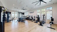 The Yardly Mount Dora fitness center at 2507 Koji St offers a well-appointed, single-floor exercise space designed to support a comprehensive workout routine. Soaring ceilings and an abundance of large, black-framed windows flood the room with natural light while offering outdoor views of palm trees and the surrounding community, creating an open and energizing atmosphere.

The light wood flooring throughout lends a clean, modern aesthetic that complements the bright white walls adorned with motivational phrases such as "Stronger Than Yesterday," "Fuel Your Flow," and "Embrace the Challenge." Overhead ceiling fans and recessed lighting ensure comfort and visibility throughout every workout.

The cardio section is equipped with a lineup of treadmills, ellipticals, stationary bikes, and a rowing machine, each featuring digital screens for performance tracking. A multi-functional cable and strength machine anchors the free weights area, which includes a full rack of dumbbells and a dedicated bench. A separate functional training zone features medicine balls, resistance bands, a BOSU ball, and a squat rack with weight plates.

Mirrored walls in the strength area enhance the sense of space, while a conveniently located water fountain and sanitation station promote hygiene and hydration. Ceiling-mounted televisions add entertainment options, rounding out a thoughtfully designed facility built for serious fitness pursuits.