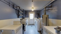 This single-floor communal laundry facility is a well-organized, functional space designed to serve multiple users efficiently. The room features rows of full-sized washing machines and dryers lining both sides, providing ample capacity for shared use. The walls are painted in a muted blue-gray tone, lending the space a clean, utilitarian aesthetic. Overhead, a central ceiling light fixture provides consistent illumination throughout the room, complemented by natural light streaming in through a white-framed exterior door at the far end, which also offers convenient access to the outdoors. The flooring is a durable, easy-to-clean surface suited for a high-traffic utility environment. Along one wall, a bank of electrical meters and circuit breaker panels is prominently displayed, reflecting the space's role as a central utility hub. A large water heater is positioned toward one end of the room, alongside various pipes and utility connections that underscore the practical infrastructure in place. The layout is straightforward and efficient, with clear pathways between the appliances for ease of movement. Overall, the space prioritizes functionality and accessibility, making it a reliable and well-equipped laundry facility.