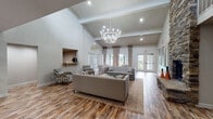Located at 5900 Old Hickory Blvd in Nashville, Tennessee, this single-story community clubhouse offers a refined and welcoming atmosphere designed for comfort and social engagement. The centerpiece of the space is a dramatic great room with soaring vaulted ceilings accented by exposed beams and an impressive statement chandelier that anchors the room with elegance. Rich hardwood-style flooring flows throughout, complementing the warm, neutral color palette of the walls and furnishings.

The main lounge area is generously proportioned, featuring tufted gray sofas, patterned accent chairs, and a stone-topped coffee table arranged atop a layered area rug — creating an inviting setting for conversation and relaxation. A floor-to-ceiling stacked stone fireplace serves as a bold architectural focal point along one wall, adding texture and warmth to the open interior.

Multiple sets of French doors line the perimeter, flooding the space with natural light and providing easy access to the outdoors. Decorative drapery panels and large-scale wall art enhance the refined aesthetic throughout. A dedicated office or business center area is visible within the open floor plan, equipped with a workstation and pendant lighting. Built-in niched shelving and a coffee station add functional charm. The overall ambiance is polished yet approachable, ideal for community gatherings.