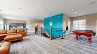 This well-appointed community space offers a generously sized, single-floor layout designed for socializing and recreation. The open lounge area is anchored by a large sectional leather sofa and flooded with natural light through multiple windows framed by soft sage-green walls, creating a welcoming and relaxed atmosphere. Patterned carpeting adds texture and visual interest throughout the main gathering areas.

A bold teal accent wall serves as a striking focal point, adding a vibrant pop of color that ties the space together. Adjacent to the lounge is a dedicated game area featuring a full-sized pool table, providing an entertaining option for guests and residents alike.

The kitchen is well-equipped for communal use, featuring rich wood cabinetry, stainless steel appliances including a refrigerator and microwave, a large white-topped island with teal bar seating, and light wood-look flooring. It is bright and functional, ideal for hosting gatherings or preparing refreshments.

The hallway connects the various areas of the floor and leads to restroom facilities, providing practical flow throughout the space. A staircase with a handsome wood-and-iron railing provides access between levels. Recessed lighting throughout ensures the space is well-lit, complementing the ample natural light that streams in from the surrounding windows.