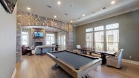 This single-level clubroom blends rustic charm with modern amenities, creating a welcoming and versatile gathering space. The centerpiece of the room is a striking herringbone-patterned brick archway that separates the lounge area from the game room, adding architectural character throughout. The lounge area is anchored by a floor-to-ceiling brick fireplace topped with a wall-mounted TV, complemented by comfortable seating including a neutral sofa, leather armchairs, and a solid wood coffee table — ideal for relaxed socializing. Crown molding and recessed lighting run throughout, lending a polished finish to the warm, neutral-toned walls. The game area features a full-size pool table and a large farm-style dining table, both bathed in abundant natural light streaming through tall windows with decorative transoms that frame outdoor views. A separate kitchen area is outfitted with dark espresso cabinetry, granite countertops, stainless steel appliances including a gas range and refrigerator, and a brick-accented island with casual seating — offering full functionality for hosting events. Light hardwood flooring flows seamlessly throughout the space, tying together the rustic and refined design elements that define this well-appointed clubroom.