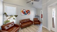 Located at 700 Moraine Ct in Virginia Beach, VA, this single-floor commercial space offers a thoughtfully designed layout comprising an entrance and two distinct living room areas. The interior features light gray walls, crown molding, and light-toned hardwood-style flooring throughout, creating a clean, cohesive aesthetic.

The entrance area sets a professional tone with frosted glass French doors that divide the space while allowing natural light to flow freely between rooms. A whitewashed vanity with a mirror provides a functional yet elegant touch, ideal for a client-facing setting.

The first living room serves as a well-appointed waiting or reception area, furnished with a tufted cognac leather sofa and matching armchairs that add warmth against the neutral palette. Vaulted ceilings with a ceiling fan, multiple windows with blinds, and vibrant abstract wall art contribute to an inviting and lively atmosphere. A potted tropical plant adds a natural accent.

The second living room functions as an amenities or refreshment area, equipped with a wall-mounted flat-screen TV, a water dispenser, a compact beverage refrigerator, a coffee station, and a cane-front cabinet — all designed to enhance the visitor experience.

Together, the rooms create a polished, comfortable environment well-suited for a professional office, wellness practice, or boutique studio in Virginia Beach.