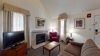 Located at 284 Daniel Webster Highway in Merrimack, New Hampshire, this two-floor suite is thoughtfully designed to accommodate both short and extended stays. The layout spans two levels connected by a staircase, offering a clear separation between living and sleeping areas.

The main level centers on a carpeted living room furnished with a striped sofa, armchairs, and a coffee table arranged around a tile-surround fireplace — a standout feature that adds warmth and character to the space. Natural light filters in through curtained windows, and a flat-screen television on a wooden dresser completes the sitting area. Adjacent to the living room is a compact, fully equipped kitchen featuring dark wood cabinetry, a full range with oven, sink, and countertop space — practical for meal preparation during longer visits.

Upstairs, the bedroom provides a quiet retreat with a queen bed, wooden headboard, nightstand, and a dedicated work desk with an ergonomic chair positioned near the window. A closet and dresser offer ample storage. The bathroom is finished with tile flooring, a tub/shower combination, wood-toned vanity cabinetry, and a large mirror with overhead lighting. Vaulted ceiling lines on the upper level lend an open, airy quality to the space overall.