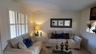 Located in Euless, Texas, this single-floor residence offers a comfortable and thoughtfully arranged living environment. The living room serves as the heart of the home, showcasing a warm and inviting ambiance achieved through a soft, neutral color palette on the walls and light carpeted flooring. Plush cream sofas are styled with decorative accent pillows in rich tones of teal and deep brown, adding depth and personality to the space. A glass-top coffee table anchors the seating area, adorned with elegant decorative accents. Warm lamp lighting creates a cozy atmosphere, complementing the natural light that filters in through window blinds. A framed triptych wall art piece adds a refined, finished touch to the main wall. The overall design of the living area reflects a balance of comfort and style, making it an ideal space for relaxing or entertaining. The single-floor layout provides convenient, easy access throughout the home, contributing to a functional and seamless flow between spaces. This residence in Euless offers a welcoming setting within a well-established Texas community.