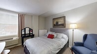 Located at 123 N Broad St in Lancaster, Ohio, this single-floor space offers a practical and efficiently arranged layout comprising one bedroom, one bathroom, and a closet. The bedroom serves as the primary living area and occupies the majority of the total floor space, providing ample room for a full-sized bed, a vanity with mirror, a multi-drawer dresser, and a seating area with a cushioned armchair. Neutral gray walls and carpet flooring lend a calm, understated aesthetic throughout. A framed landscape painting with a decorative gold frame anchors the bedroom wall, adding a touch of classic character. A floor lamp provides warm ambient lighting, complementing the natural light that enters through a window fitted with blinds and curtain panels. A compact workspace area near the entry features a desk with a television and microwave, highlighting the room's functional versatility. The bathroom is efficiently appointed with a full-sized soaking tub and shower combination, chrome fixtures, a grab bar for added safety, and glass shelving for storage. A closet offers additional organizational space. The bold black entry door adds a distinctive architectural contrast to the otherwise light interior palette, giving the space a memorable first impression.
