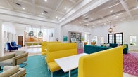 Located at 650 Dekalb St in Auburn, Alabama, this expansive single-floor community space offers a thoughtfully designed interior that blends classic architectural details with a bold, contemporary aesthetic. The space is anchored by striking coffered ceilings throughout, lending a sense of height and grandeur to the open layout. Light hardwood flooring flows seamlessly across the entry and main areas, while distinct zones are defined by vibrant area rugs in teal and magenta.

The entry welcomes visitors with soaring ceilings, a decorative gold-framed mirror, and dark-framed French doors that flood the space with natural light and offer views to the outdoors. The main lounge area is centered around a floor-to-ceiling natural stone fireplace, above which a flat-screen television is mounted. Plush teal sofas, gold wingback chairs, and colorful accent pillows contribute to the lively, energetic atmosphere.

Adjacent seating zones feature bold yellow booth-style seating paired with white tables, ideal for collaborative gatherings. A sunlit lounge area lined with large windows and French doors overlooks the surrounding greenery, creating an airy, inviting retreat. Built-in shelving displays curated décor, and recessed lighting complements the natural light throughout. A decorative chandelier adds an artistic focal point near the entry, completing this vibrant and well-appointed space.