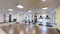 This single-floor fitness center offers a clean, modern environment designed to support a well-rounded workout routine. The space is outfitted with a variety of cardio equipment, including multiple treadmills and elliptical machines, each equipped with individual screens for entertainment during workouts. A dedicated free weights station with a dumbbell rack is positioned along one wall, providing options for strength training. The flooring transitions from rich hardwood in the open exercise areas to carpet beneath the cardio equipment, offering both durability and comfort underfoot. Bright, recessed panel lighting in the ceiling ensures consistent illumination throughout, while ceiling fans contribute to air circulation and comfort. Large windows along the far wall allow natural light to fill the space and provide views of the outdoors, creating an open and energizing atmosphere. The walls are finished in neutral tones, keeping the environment clean and focused. A shelving unit offers additional storage for equipment or personal items. The overall layout is thoughtfully organized, providing ample room to move freely between stations, making this fitness center a functional and inviting space for daily exercise.
