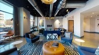 Located at 104 Washington St in downtown Dover, NH, this single-floor coworking space delivers a contemporary, design-forward work environment. The open lounge area serves as the heart of the space, featuring exposed ductwork against a dramatic black ceiling, bold geometric rugs, curated artwork, and eclectic seating including navy sofas, leather ottomans, and accent chairs arranged around a modern fireplace. Floor-to-ceiling windows flood seating nooks with natural light and offer street-level views of the surrounding urban landscape, with counter-height window seating ideal for focused work. Glass-partitioned private offices and phone booths provide acoustic privacy while maintaining visual openness, and multiple conference rooms are outfitted with large wooden tables, teal barrel chairs, integrated AV technology, and whiteboard walls for collaborative sessions. The polished concrete floors and teal accent baseboards create a cohesive industrial-chic aesthetic throughout the corridors. A well-appointed kitchen features bold emerald green tile, open stainless steel shelving, a coffee station, a beverage cooler, and a full sink. A dedicated fitness center with glass-wall access rounds out the amenities, offering a comprehensive and functional workspace experience.