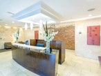 Located at 90–98 Sterling Blvd in Englewood, NJ, this single-floor lobby space presents a well-appointed, professional environment designed to welcome and orient visitors. The interior is defined by large-format cream travertine tile flooring that extends throughout, complemented by matching stone-clad walls that lend a cohesive, upscale aesthetic.

At the heart of the space sits a substantial curved reception desk finished in rich dark wood with a light stone countertop. Pendant lighting and a recessed tray ceiling with integrated downlights illuminate the desk area, creating a clear focal point. Decorative floral arrangements add a refined, inviting touch.

Near the secured entry, a seating alcove features two leather armchairs positioned on a striped area rug beneath warm lamp lighting, offering a comfortable waiting area. Black-framed glass doors and partitions provide secure access points while maintaining visual openness.

Recessed can lighting throughout ensures consistent, even illumination across the space. An adjacent room, visible through an open doorway, reveals hardwood flooring, a kitchen-style counter with bar seating, and an exposed stacked-stone accent wall, suggesting a resident or staff amenity area. The overall ambiance is polished, functional, and professionally maintained.