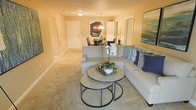 This single-floor residence offers a thoughtfully designed open-concept layout that seamlessly connects the main living and dining areas. The living room is a welcoming, light-filled space anchored by a generously sized sectional sofa adorned with navy and teal accent pillows, paired with a set of round nesting coffee tables that add a contemporary touch. Soft, neutral-toned carpeting runs throughout, contributing to the warm and comfortable ambiance. Warm beige walls and flush-mount ceiling lighting create an inviting glow across the living area. A striking large-scale artwork adds a bold focal point to the room. Transitioning toward the back, the dining area features an elegant chandelier suspended above a dark dining set, lending a touch of sophistication. Multiple doorways lead to additional rooms, suggesting a well-organized and functional floor plan. The overall aesthetic is clean and modern with neutral tones punctuated by coordinated accent décor, making this space both stylish and livable. The open sightlines between the living and dining zones enhance the sense of spaciousness throughout the home.