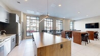 Located at 70 Park East Dr in Winnipeg, Manitoba, this single-floor communal amenity space offers a well-appointed and contemporary environment designed for shared use. The open-concept layout is anchored by a fully equipped kitchen featuring sleek grey cabinetry, a textured dark tile backsplash, stainless steel appliances including a full-size refrigerator and range, and a generous wood-topped island with built-in bar seating. Pendant lighting runs above the kitchen area, providing focused illumination that complements the recessed ceiling lights throughout.

The adjacent lounge and dining area is thoughtfully furnished with multiple high-top tables in warm wood tones paired with black bar stools, encouraging a casual and social atmosphere. A built-in banquette with white upholstery and small pedestal tables offers an additional intimate seating option, accented by sleek pendant fixtures above.

Large floor-to-ceiling windows with black frames line the perimeter, flooding the space with natural light and offering views of the surrounding Winnipeg streetscape. Light-toned large-format tile flooring runs seamlessly throughout, enhancing the clean, bright aesthetic. The overall design balances modern functionality with an inviting, communal ambiance suited to a residential amenity setting.