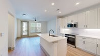 This thoughtfully designed 1-bedroom, 1-bathroom residence offers a clean, modern aesthetic across a single floor. Light-toned hardwood-style flooring flows consistently throughout the space, unifying each room with a warm, cohesive feel.

The open-concept kitchen and living area serves as the heart of the unit. The kitchen is well-appointed with white shaker-style cabinetry extending to the ceiling, stainless steel appliances including a French door refrigerator and range with microwave, and a large central island featuring a single-basin sink with a matte black faucet and ample quartz countertop space. Recessed lighting illuminates the area efficiently.

The bedroom is a comfortable, well-proportioned room benefiting from large windows that invite natural light and outdoor views. A ceiling fan adds both function and style. Interior doors feature a modern paneled design with matte black hardware, consistent with the unit's overall design language.

The bathroom is finished with a white vanity, quartz countertop, undermount sink, and matte black fixtures. A glass-enclosed walk-in shower with a tiled surround adds a refined touch. Directly accessible from the bathroom is a dedicated walk-in closet with built-in shelving and hanging rods, providing practical storage.

A hallway connects the rooms efficiently, completing this well-planned, move-in-ready layout.