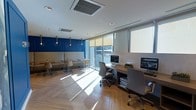 This single-floor office space combines functionality with contemporary design across two distinct zones. The workstation area features a long wooden desk equipped with desktop computers and ergonomic task chairs, positioned along floor-to-ceiling windows fitted with roller blinds. These expansive glazed panels allow generous natural light to flood the light wood herringbone flooring, creating an open and productive atmosphere.

Adjacent to the workstation zone, a casual lounge or breakout area is defined by a striking deep navy blue accent wall with classic paneling detail. This section includes a neutral upholstered bench seat, small bistro-style tables, and modern shell chairs, offering an informal setting for meetings or relaxation. Pendant lighting with warm-toned brass fixtures hangs above this area, adding an elegant contrast to the bold wall color.

The ceiling features recessed LED downlights and a centrally mounted cassette-style air conditioning unit, ensuring consistent climate control throughout. A glazed sliding door or large window panel connects the interior to what appears to be an exterior balcony or terrace, further enhancing the sense of openness and ventilation within the space.
