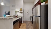 Located at 2025 Ayrsley Town Blvd in Charlotte, NC, this one-bedroom, one-bathroom residence offers a thoughtfully designed layout that maximizes every inch of its compact footprint. Soaring ceilings throughout lend an open, airy quality to the space, making the living areas feel more expansive than their physical dimensions suggest.

The galley-style kitchen is well-equipped with dark espresso cabinetry, granite countertops, stainless steel appliances including a full-size refrigerator, dishwasher, and range, and open floating shelves for additional storage. Hardwood-style plank flooring runs throughout the kitchen and living areas, adding warmth and continuity.

The living room receives natural light through a large window overlooking the surrounding neighborhood, creating a bright and inviting atmosphere. Neutral wall tones complement the space and provide a versatile backdrop.

The bedroom features plush carpet, a double-door closet, and a window with views of the Ayrsley community. High ceilings continue here, reinforcing the sense of openness.

The bathroom is finished with matching granite countertops, dark wood cabinetry, tile flooring, and open shelving, maintaining the design consistency found throughout the residence.